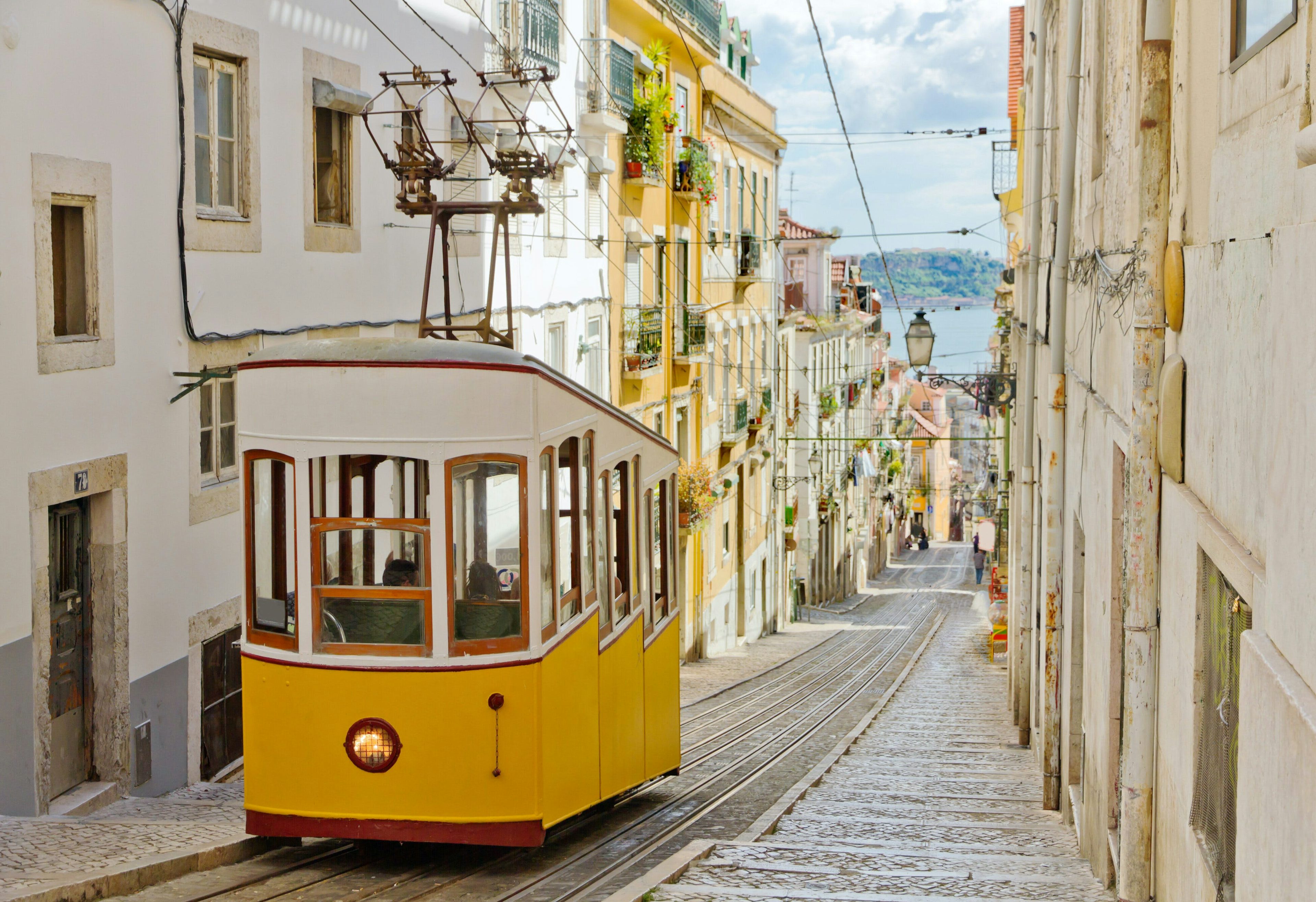 Yellow streetcar ascending a steep cobblestone street in Lisbon, with historic buildings lining the sides.