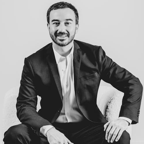 Black and white portrait of James Muscat, CEO and Founder of Moviinn, seated with his left hand resting on his knee and his right arm draped over his other leg. He is dressed in an elegant dark suit complemented by a crisp white shirt, smiling, exuding a professional and friendly look.