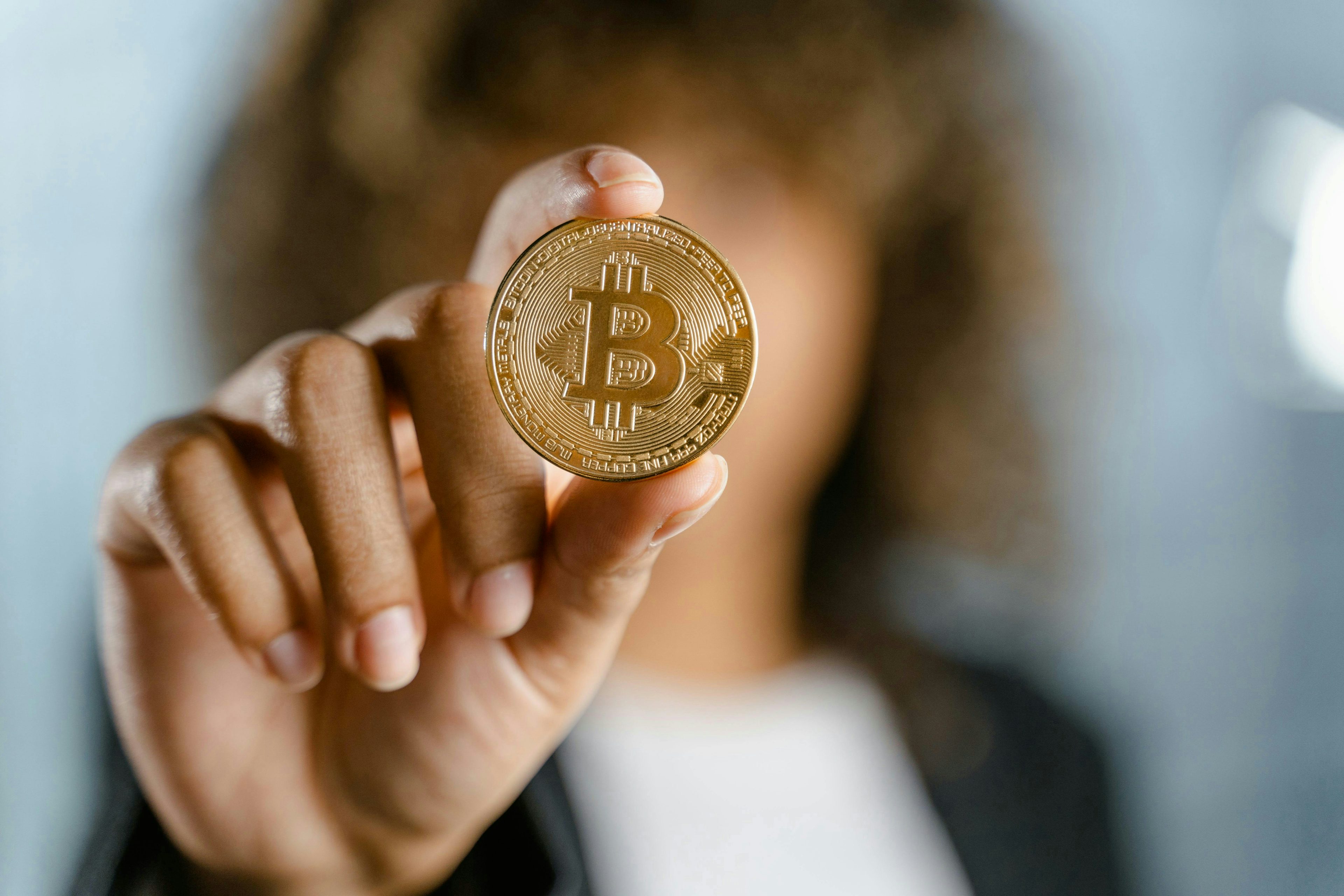 Uma imagem aproximada de uma moeda Bitcoin exibida primeiro plano, segurada por uma mulher com a mão em foco, com uma imagem desfocada dela ao fundo.
