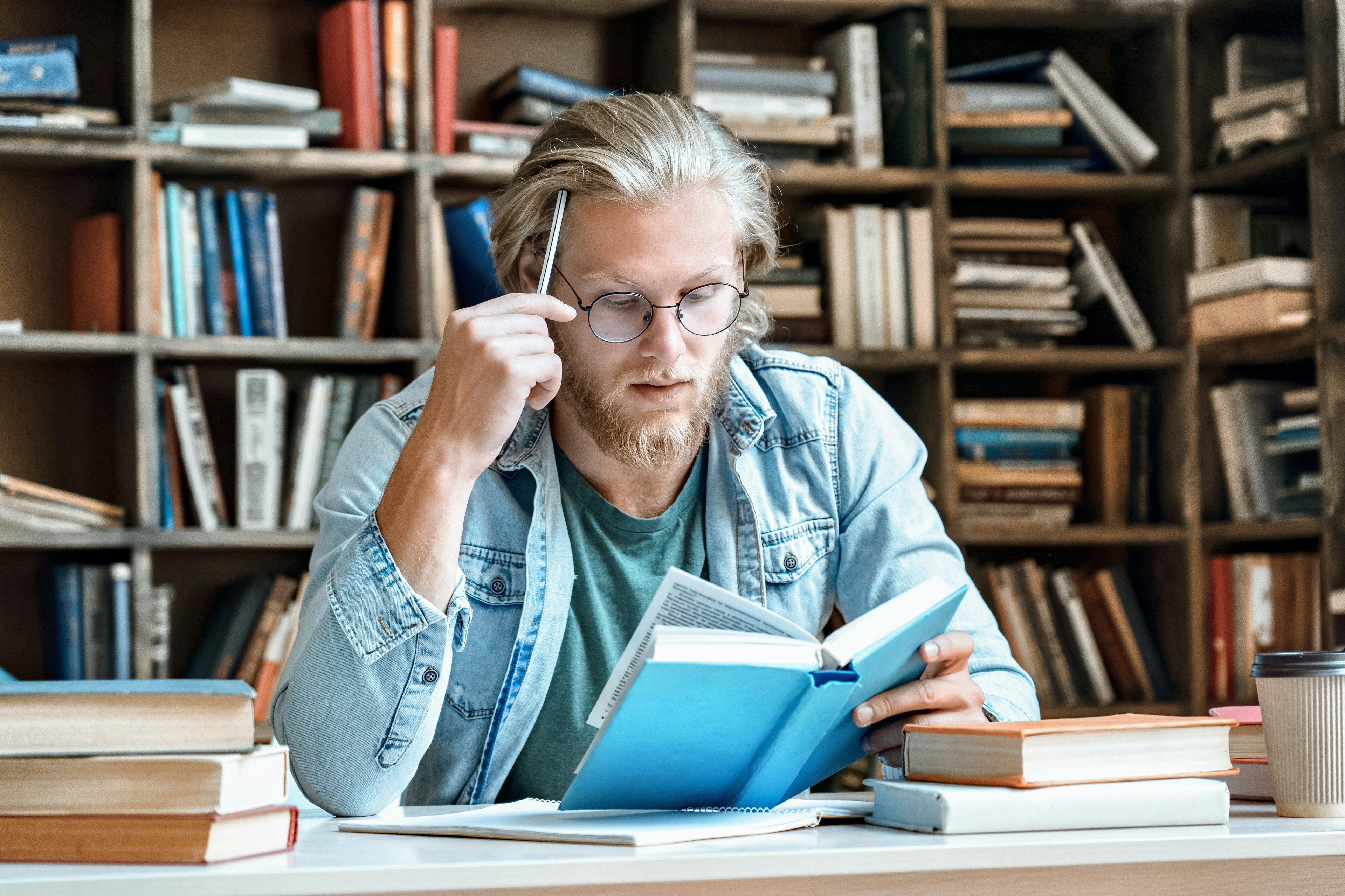 Blond man with round glasses holding a pencil against his forehead while intently reading a book.