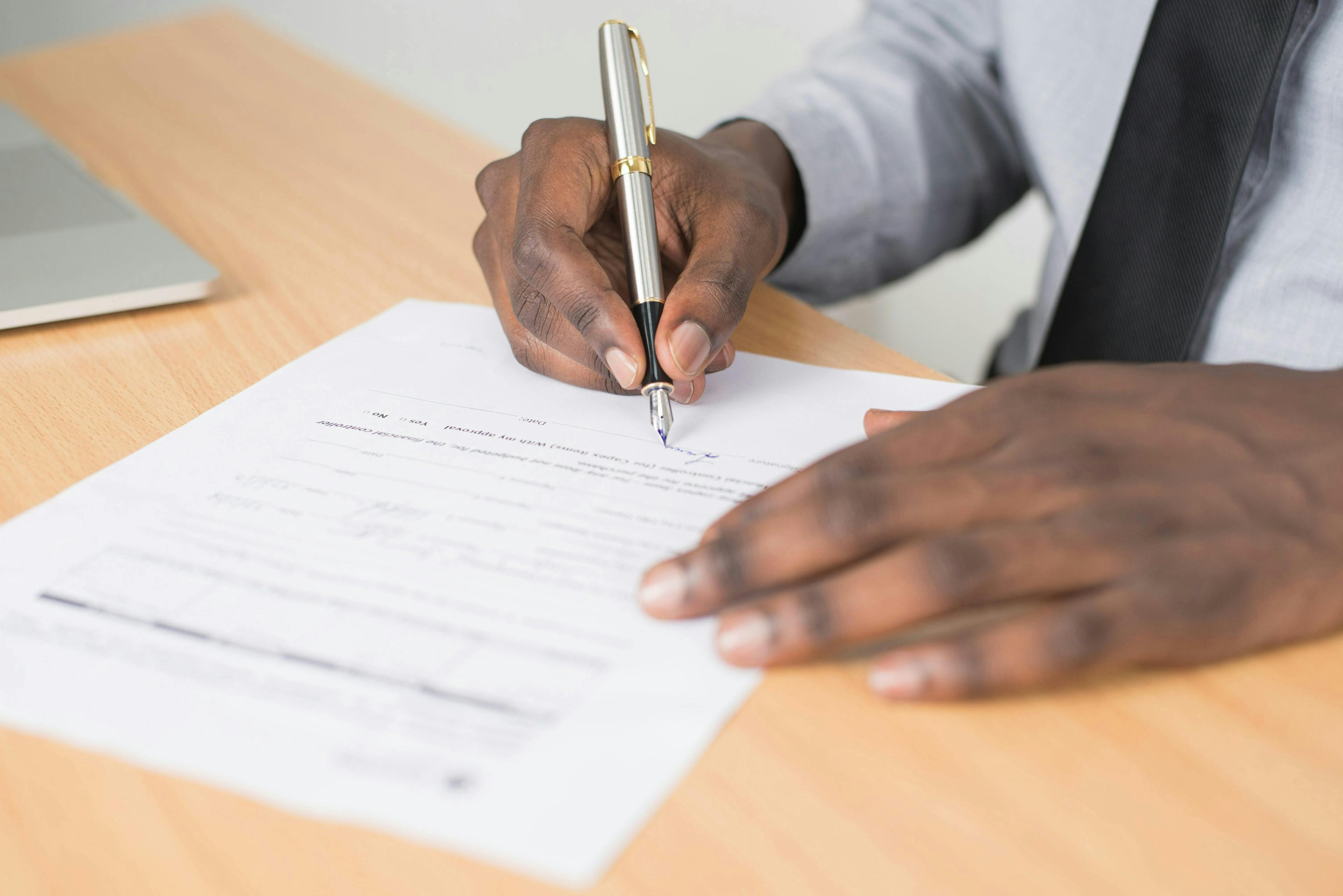 A close-up image showing only the hands of a man as he signs an application form. One hand is gently resting on the form, providing stability, while the other holds a sleek silver pen, poised in the act of signing. The focus is on the precise movement of the pen on the paper.