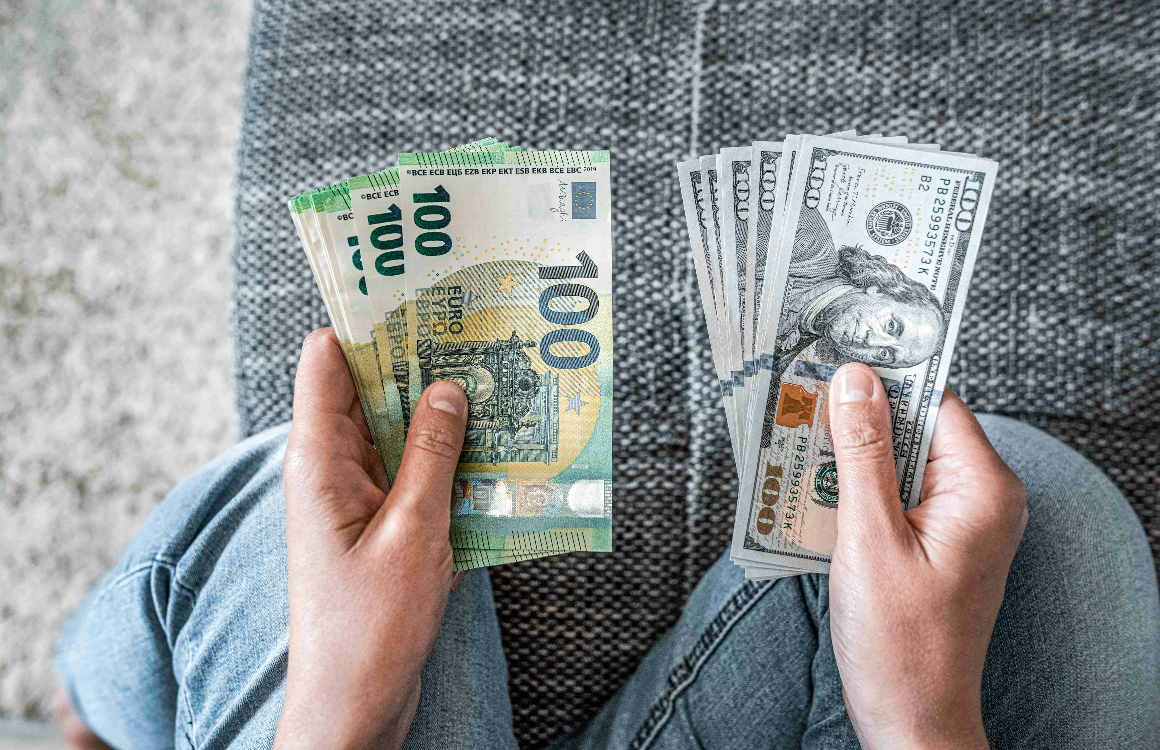 Pessoa sentada de calça jeans, segurando notas de euro na mão esquerda e notas de dólar americano na mão direita.